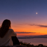 Woman seated overlooking the water at sunset, gazing at a bright point of light in the twilight sky.